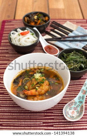 Asian soup with shrimps in white ceramic bowl composed with ceramic spoon with spicy red sauce, asian chopsticks and black bowls with rice noodles, kale (green cabbage) and fried vegetables.