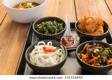 Asian dish on a square plate in black bowls with shrimp, rice noodles, kale (green cabbage) and fried vegetables. Composed with ceramic spoon with spicy sauce, chopsticks and a bowl of asian soup.