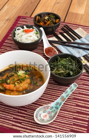 Japanese soup with shrimps in white ceramic bowl composed with ceramic spoon with spicy red sauce, japanese chopsticks and black bowls with rice noodles, kale (green cabbage) and fried vegetables.
