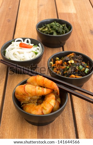 Asian vegetarian food composed with four black bowls with shrimps, rice noodles, kale (green cabbage), fried vegetables and chinese chopsticks. Composition on a old styled wooden table.