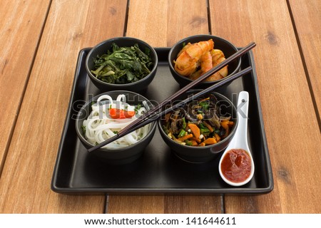 Asian dish on a square plate in black bowls with shrimp, rice noodles, kale (green cabbage) and fried vegetables. Composed with ceramic spoon with spicy red sauce and chinese chopsticks.