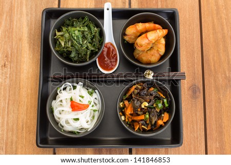 Traditional asian dish on a square plate in black bowls with shrimp, rice noodles, kale (green cabbage) and fried vegetables. Composed with ceramic spoon with spicy red sauce and chinese chopsticks.