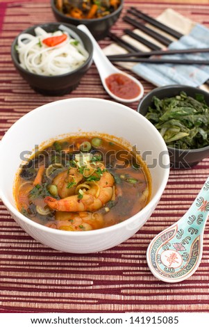 Chinese soup with shrimps in white ceramic bowl composed with ceramic spoon with spicy red sauce, asian chopsticks and black bowls with rice noodles, kale (green cabbage) and fried vegetables.