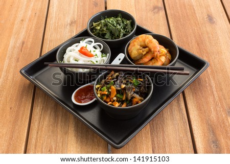 Traditional japanese dish on a square plate in black bowls with shrimp, rice noodles, kale (green cabbage) and fried vegetables. Composed with ceramic spoon with spicy red sauce and chinese chopsticks