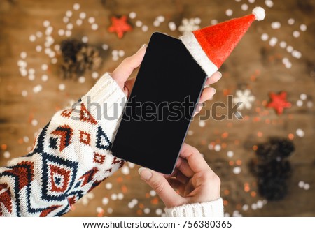 Female hands holding smart mobile phone with oled display on wooden background with Christmas gifts snowflakes and snow. Happy New Year, hat santa on mobile phone, Flat lay composition top view.