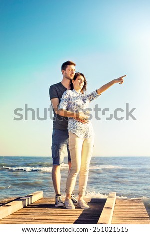 Happy young couple standing arm in arm pointing into the sky towards the hot summer sun, upper body against a clear blue sky