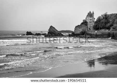 Rocky black and white, coastal beach seascape in Biarrtiz, France on the Atlantic ocean. Surfers in the water surfing waves towards a sandy beach. / Seascape in Biarrtiz, France.