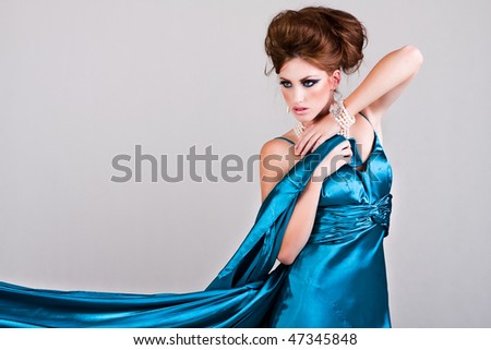 Attractive young woman standing in a blue satin dress that is being blown in the wind. Horizontal shot.