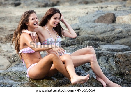 Two attractive young women sit on a rocky shore with their arms around each other. Horizontal shot.