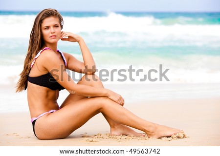 An attractive young woman wearing a black bikini sits on a beach with her elbow on her knee and her hand to her head.  Surf can be seen in the background. Horizontal shot.