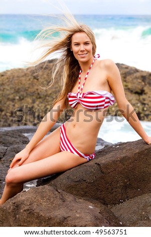 An attractive young woman sits on rocks at the beach with her hand on her leg. She is wearing a red and white striped bikini. Vertical shot.