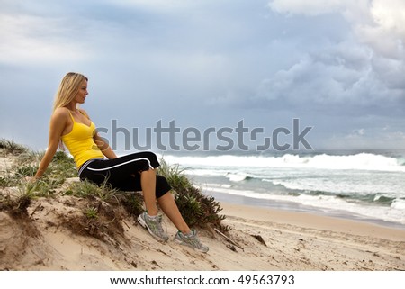 Full length profile portrait of a young woman sitting on a grassy bluff on the beach. She is staring out at the sea with a serious expression. Horizontal shot.