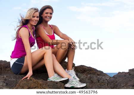 Low angle portrait of two young women sitting on rocks at the beach. They are wearing tank tops and shorts and smiling at the camera. Horizontal shot.