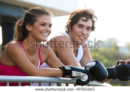 Young couple leaning over a railing in an outdoor setting. They are both wearing boxing gloves and smiling. Horizontal shot.