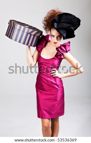 A young woman dressed in avant garde attire and carrying a hat box. She is wearing a hat and has cosmetic artwork on her right temple. Vertical shot.