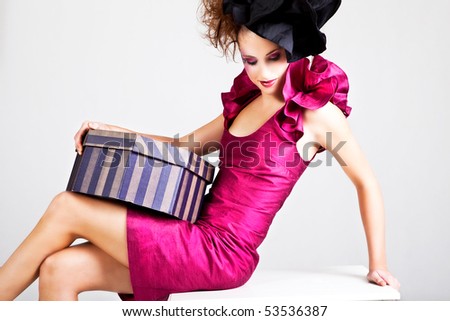 A young woman dressed in avant garde attire and holding a hat box. She is wearing a hat and has cosmetic artwork on her right temple. Horizontal shot.