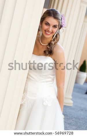 An attractive young woman wearing a white dress and pearls is smiling and standing behind a pillar. She has a purple flower in her hair.  Vertical shot.