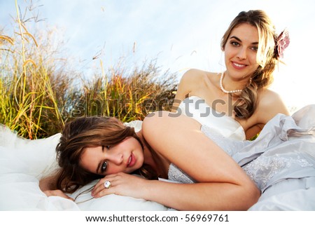 An attractive young bride and her bridesmaid are lying in a grass field. The bridesmaid is lying across the lap of the bride. Horizontal shot.