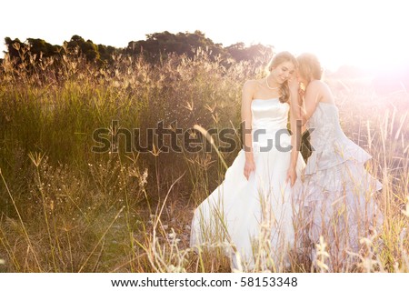 Attractive young bride and bridesmaid in formal attire. They are walking in the country holding hands at sunset.