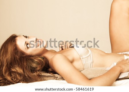 Sensual young woman with long red hair lies atop pillows. She is wearing a strapless bra and smiling upwards. Horizontal shot.