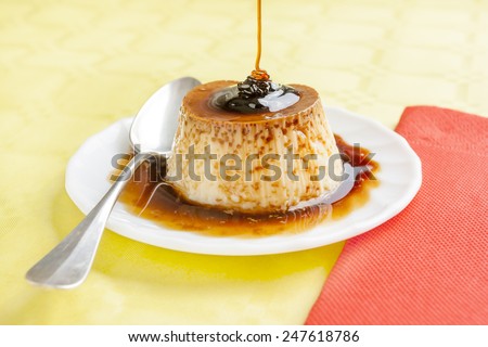 Pouring syrup on vanilla flan with red napkins and spoon on yellow table