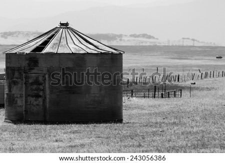 Black and white image of a rusty crop storage bin with broken fence and mountains in background