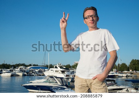 Man showing victory gesture of two fingers on the background bay and yachts in pretty summer day.