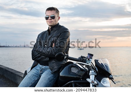 Handsome young man in leather jacket on motorcycle on sunset light on the sea embankment.