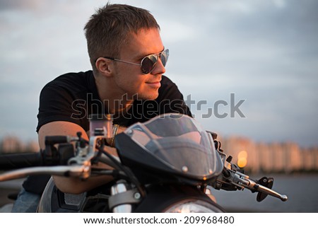 Romantic portrait handsome  biker man in sunglasses sits on a bike on a sunset near lake and city