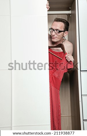 curious naked man in eyeglasses looking out from white wardrobe hide behind the woman\'s red dress in white dots.