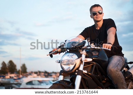 Romantic portrait handsome biker man in sunglasses sits on a bike on a sunset near lake and yacht club. copy space left