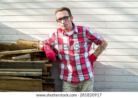 Man in casual clothes and rubber working gloves near his country house leaning on stack of wood.