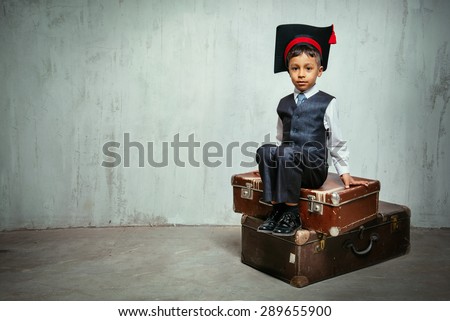 small black boy in suit and graduation hat sits on the old suitcases and look into the camera. copy space left, instagram toned