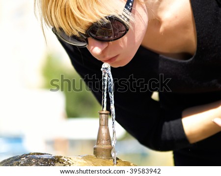 The woman drinks water from a small fountain in the summer in the street