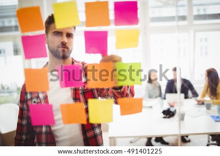 Photo editor looking at multi colored sticky notes on glass in meeting room at creative office