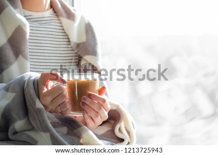Woman's hands holding hot steaming drink. Female covered in throw blanket sits by the window with cup of hot cocoa