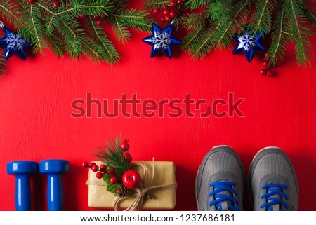 Christmas sport flat lay composition with blue dumbbells sneakers gift box and spruce tree branches red wooden background. Concept Christmas special for healthy lifestyle and sport.