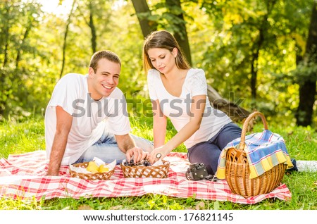 Happy young couple having a picnic eating grapes and sitting on the picnic cloth enjoying the autumn nature in the park