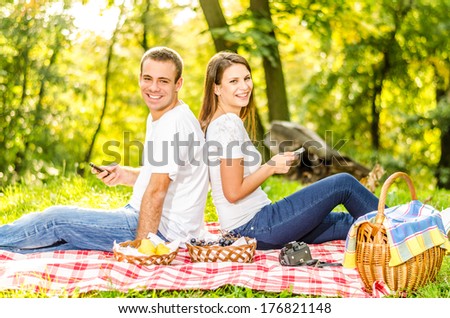 Happy young couple having a picnic typing the mobile and sitting on the picnic cloth enjoying the autumn nature in the park