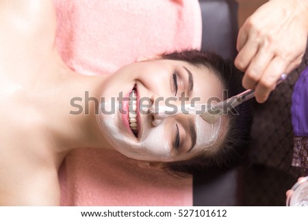 people, beauty, spa, cosmetology and skincare concept - close up of beautiful young woman lying with closed eyes and cosmetologist applying facial mask by brush in spa center