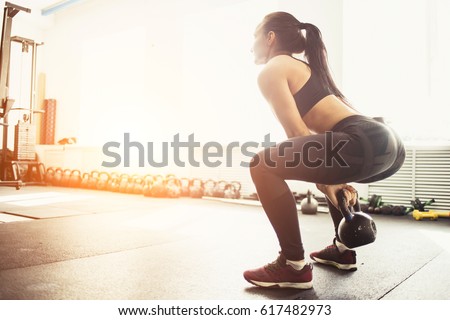 Athletic woman exercising with kettle bell while being in squat position. Muscular woman doing cross fit workout at gym.