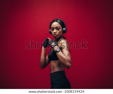 African young woman wearing boxing gloves posing in combat stance looking at camera. Fit young female boxer with headphones ready for fight on red background.