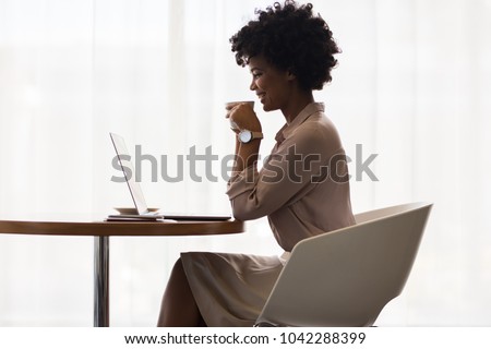 African businesswoman smiling while using laptop and drinking coffee in office. Side view of office worker having coffee while working on laptop.