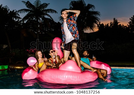Man standing on inflatable mattress and looking forward with friends sitting at back. Group of men and women partying in the pool at night.