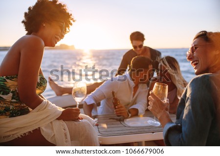 Group of men and women having a boat party during sunset. Happy young friends sitting at the back of a yacht and having fun.