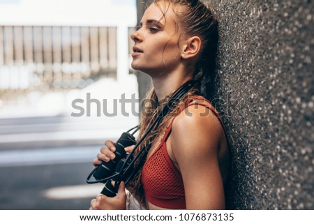 Tired and exhausted woman resting after intense training session. Female with skipping rope leaning to a wall and taking break from physical training.
