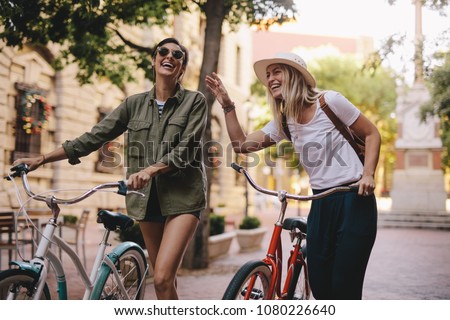 Positive and happy girls walking on the city street with bicycles. Female friends enjoying a walking down the street with their bikes.