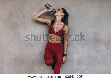 Female athlete drinking water while standing by a grey wall. Slim woman in sportswear taking a break after workout.