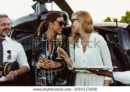 Cheerful female friends having wine outside a helicopter with pilot standing by. Women enjoying welcome drinks by a helicopter.