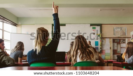 Rear view of female student sitting in the class and raising hand up to ask question during lecture. High school student raises hand and asks lecturer a question.